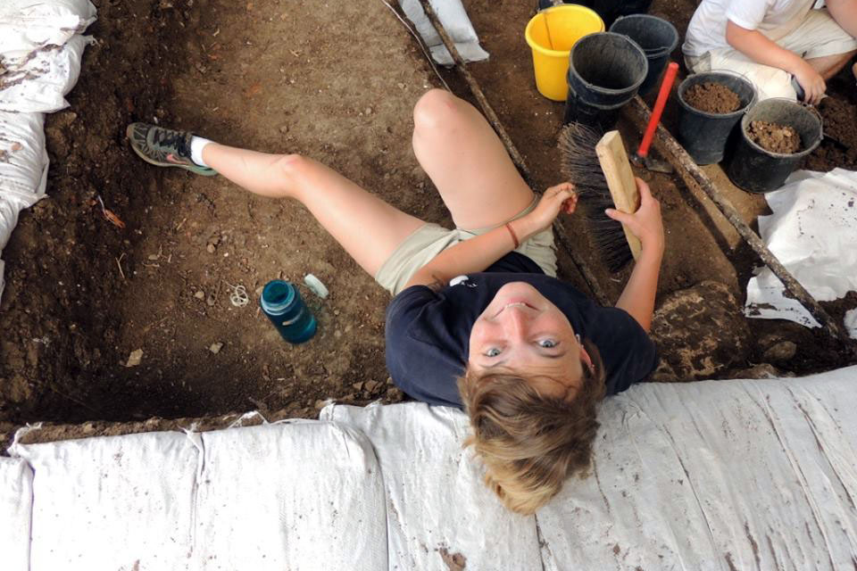 A student shown from above holding excavation materials at a dig site surrounded by dirt